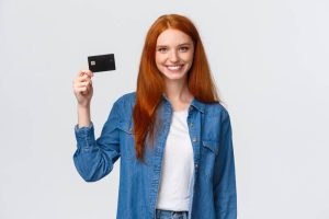 female red head in blue jean jacket and white shirt smiling holding first credit card against white background